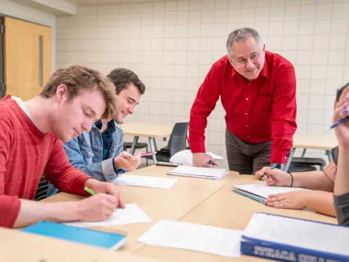 A faculty member in a red shirt is standing at a table speaking to four students seated around the table.