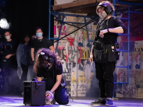 A Stage Management student dressed in all black stands next to another Stage Management student crouching down. The crouching student is taping a mark on the set of RENT.