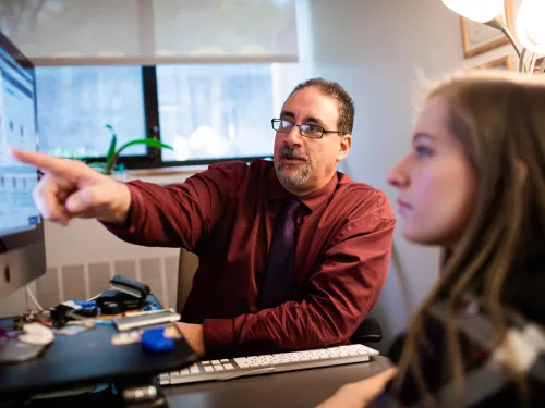 Man points to something on a computer screen, pointing it out to a female student