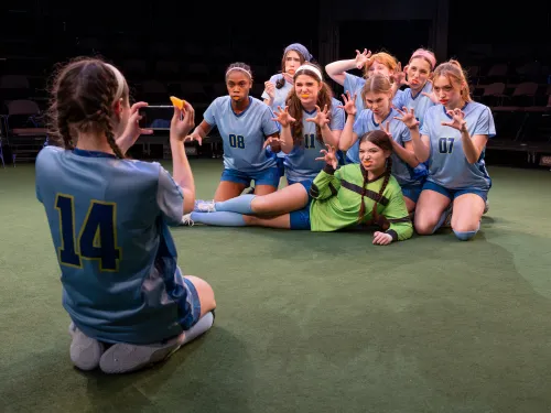 A group of female-identifying actors in blue soccer uniforms. They have orange slices in their mouths and are posing for a camera and making "wolf" expressions with their mouths and hands. Another actor is taking their picture with a cell phone.
