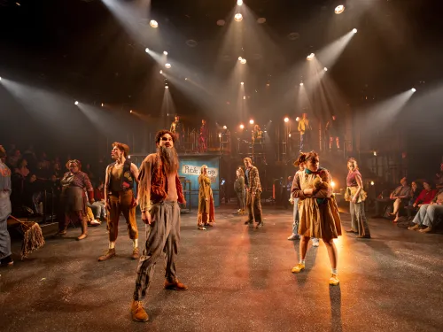 A group of actors in Urinetown The Musical standing on stage looking out. Bright yellow lights flood the stage. 