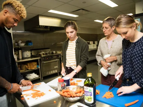 A group of students cut vegetables under the watchful eye of their professor