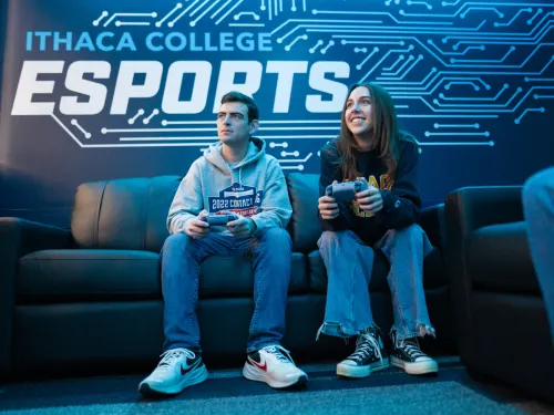 Two college students play video games on a sofa. The wall behind them says "Ithaca College Esports."