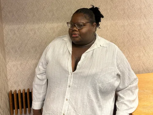 Image of Ashanti Ford '24. Ashanti is wearing a white button shirt and glasses. Ashanti is posed on a textured neutral colored wall.