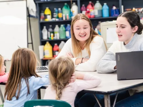two female college students sit and talk with a group of three young elementary students 
