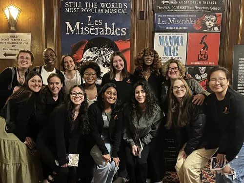 A group photo of 14 women in front of a Broadway theatre advertising Les Miserables