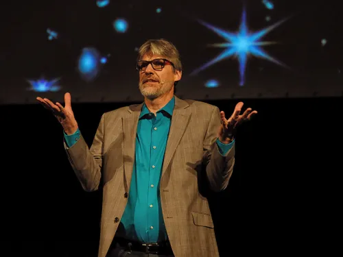 A man wearing glasses, blue shirt, and a blazer stands onstage with stars projected behind him. 