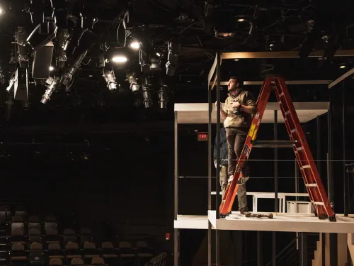 A TPD student stands on a ladder which sits on a platform. He is holding a drill and looking at lights.
