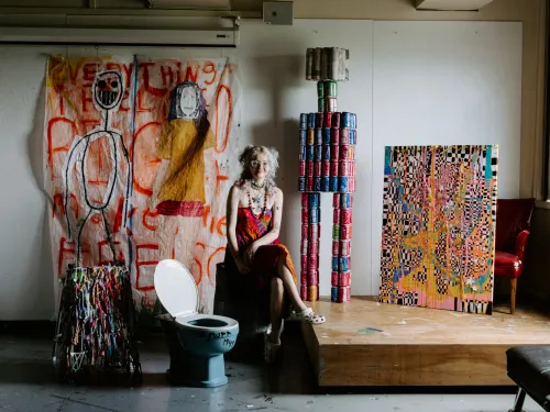 A student in an art studio is surrounded by their the objects in their installation, including a giant robot man made from discarded soda cans, a toilet, and original artwork. 