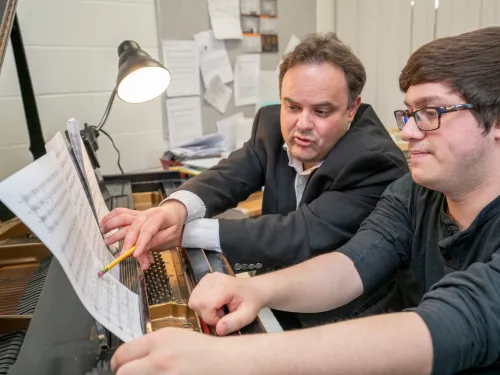 A professor and student sit side by side on a piano bench making notations on sheet music