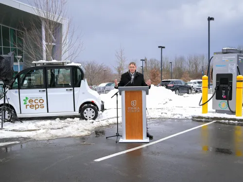 A man stands at a podium on a snowy day between an EV and an EV charger station.