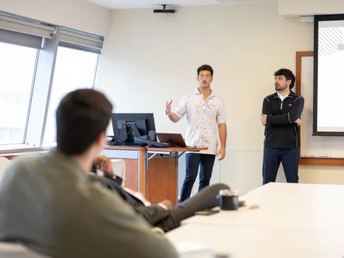 two male students stand at the front of the class giving a presentation 