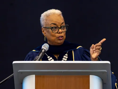A woman wearing academic regalia speaks at a podium.