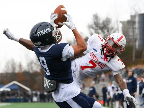 A college football player in a blue uniform catches a pass against a defender wearing a red uniform.