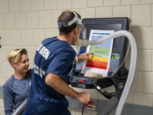 Student conducting a vo2 max test with client. The client is running on a treadmill, pointing at a graph to indicate his level of exertion for the student, to his left.