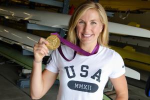 Meghan Musnicki holds up the Olympic gold medal around her neck while standing in a boathouse.