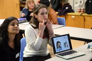 Two students are working with a client. There is a laptop computer that indicates facial expressions and the difference between the word "sick" and "seek." One student in a white sweater has her hands to her face, showing the client how to move their mouth properly to make the sounds distinctly.