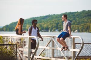 Three students are on a dock at Cayuga Lake. Two students are standing and one is sitting on the railing. It is a beautiful sunny day.