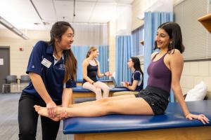 A client is seated on a physical therapy table at Ithaca College. A PT student at the end of the table is treating the patient's ankles.