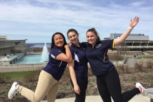 Three HSHP students are jumping up in the air excited on a spring day. In the background is the lake and a blue sky with white clouds.