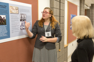 A student standing at a research poster presenting her research to a person in a hallway
