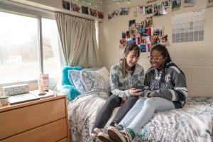 two students sitting on a bed near a window in a dorm room