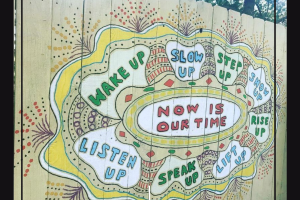 A mural painted on a light yellow fence.  The middle is a oval that reads "Now is our time" and is surrounded by bubbles that read, "wake up", "slow up", "step up", "show up", "rise up", "lift up", "speak up", and "listen up".  The outsides of the mural are decorated with sprays of dots and borders in white, yellow, and red.