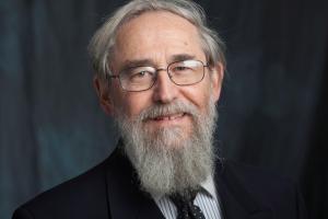 A picture of an older White man smiling at the camera.  He is wearing glasses and has a medium length grey beard that matches his grey hair.  He is wearing a dark suit and sits in front of a dark blue drop cloth.
