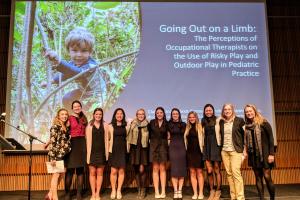 group of students stand in front of a projected slide that reads Going Out on a Limb: The perceptions of occupational therapists on the use of risky play and outdoor play in pediatric practice and features a photo of a child climbing in a tree