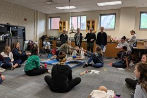 Students sit on floor and stand around a swing with a child sized mannequin on it