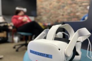 A virtual reality headset rests on a chair with a person in headset in the background
