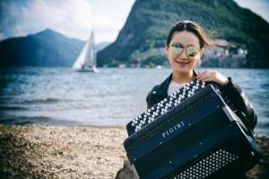A woman wearing sunglasses sits by a lake with an accordion