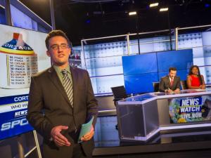 Student sportscaster speaking on set with news anchors seated in the background.