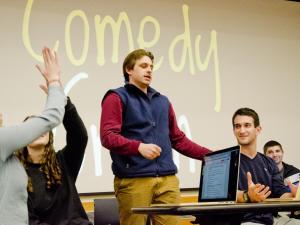 Students around open laptops discuss the topic of "comedy," written in large letters on a board behind them.