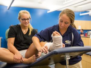 A student in a blue shirt is working on taping the ankle of another student in a blue shirt that is seated on a table.