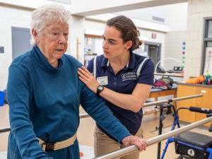 A student in a blue shirt is working with an elderly patient helping them with standing up.