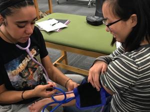 A student in a blue shirt is checking the blood pressure of another student who is seated in a chair.