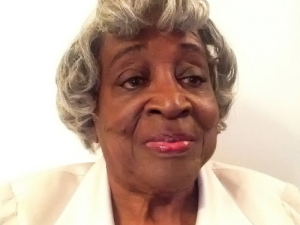 An older woman is sitting and looking slightly off to the side of the camera with a faint smile on her face.  She is wearing a white suit jacket against a white background.  Her hair ends at her ears and is salt and pepper grey.