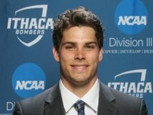 This is a photo of Jared Bauer. Jared is wearing a dark suit with a white shirt and blue striped tie. He is standing in front of a Ithaca Athletics background showing Division III athletics.