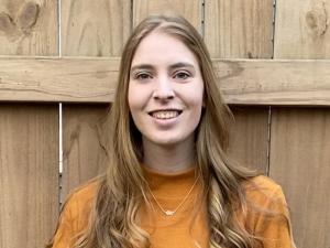 This is a photo of Paige Pickard. Paige is standing in front of a fence. Paige is wearing a long sleeve orange colored shirt.