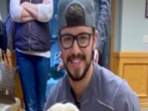 This is a photo of Zachary Thomas. Zachary is kneeling down petting a white dog. Zachary has glasses and is wearing a short sleeve blue shirt and blue jeans. Zachary is wearing a cap backwards.