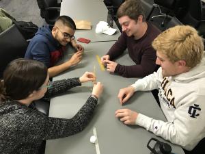 Four students sitting at a table, building a structure out of pasta.