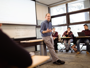 A male professor leavinging against a desk lecturing to a classroom full of students