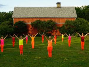 A group of dancers in brightly colored orange, red, and pink outfits stand with their arms outstretched in a green lawn in front of a barn. 