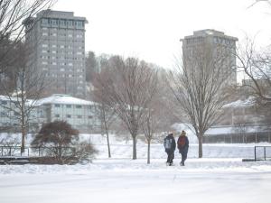Two students are walking across campus while it is snowing.