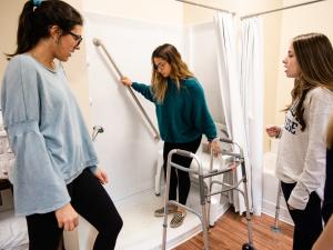 Two students are standing watching a third student in a "mock" shower, using grab bars and other assistive devices to mimic a client taking a shower.