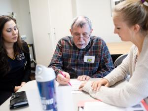 Two students are working directly with an elderly individual on writing.