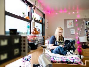 College student sitting on a bed in a dorm room with pink lights