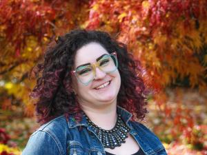 A white woman with purple hair and rainbow glasses smiles in front of fall foliage