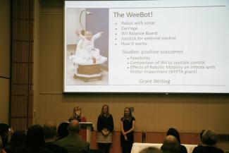 Three presenters in front of a screen showing a baby on a robot
